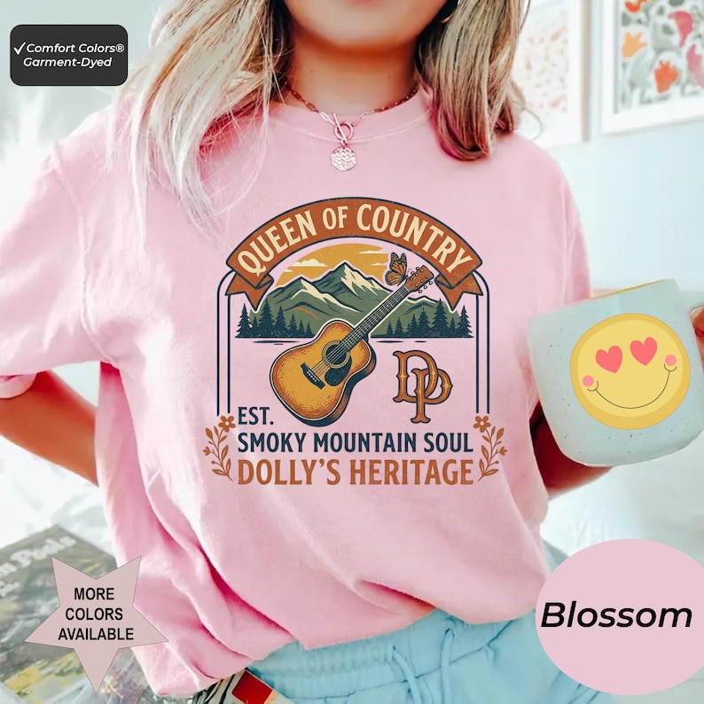 K&ouml;nnte beinhalten: Ein hellrosa T-Shirt mit einem Grafikdruck einer akustischen Gitarre, Bergen und einem Schmetterling. Der Text lautet QUEEN OF COUNTRY, EST. SMOKY MOUNTAIN SOUL, DOLLY'S HERITAGE und DP. Ein hellblauer Becher mit einem gelben Smiley wird gehalten.