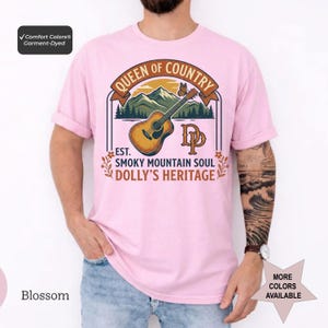 K&ouml;nnte beinhalten: Ein hellrosa T-Shirt mit kurzen &Auml;rmeln und einer Grafik aus Akustikgitarre, Bergen und Schmetterling. Der Text lautet QUEEN OF COUNTRY, EST. SMOKY MOUNTAIN SOUL, DOLLY'S HERITAGE und DP. Ein Comfort Colors Garment-Dyed Etikett ist zu sehen.