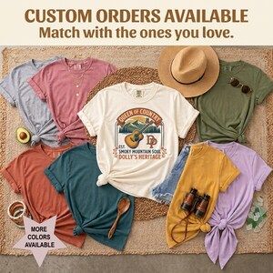 K&ouml;nnte beinhalten: Eine Sammlung von kurz&auml;rmeligen T-Shirts in verschiedenen Farben wie Creme, Grau, Rosa, Olivgr&uuml;n, Rost, Blaugr&uuml;n, Gelb und Lavendel. Das cremefarbene T-Shirt zeigt eine Grafik mit einer Gitarre, Bergen und dem Text Queen of Country.