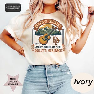 K&ouml;nnte beinhalten: Ein elfenbeinfarbenes T-Shirt mit einem Grafikdruck, der eine braune Akustikgitarre, gr&uuml;ne Berge und einen Schmetterling zeigt. Der Text lautet: QUEEN OF COUNTRY, EST. SMOKY MOUNTAIN SOUL, DOLLY'S HERITAGE und DP. Comfort Colors st&uuml;ckgef&auml;rbtes T-Shirt.