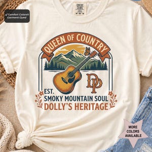 K&ouml;nnte beinhalten: Ein cremefarbenes Kurzarm-T-Shirt mit einer Grafik aus Akustikgitarre, Bergen und einem Schmetterling. Der Text lautet QUEEN OF COUNTRY, EST. SMOKY MOUNTAIN SOUL und DOLLY'S HERITAGE, erg&auml;nzt durch die Initialen DP in einer orangefarbenen Schrift.