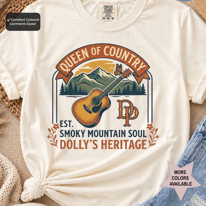 K&ouml;nnte beinhalten: Ein cremefarbenes Kurzarm-T-Shirt mit einer Grafik aus Akustikgitarre, Bergen und einem Schmetterling. Der Text lautet QUEEN OF COUNTRY, EST. SMOKY MOUNTAIN SOUL und DOLLY'S HERITAGE, erg&auml;nzt durch die Initialen DP in einer orangefarbenen Schrift.
