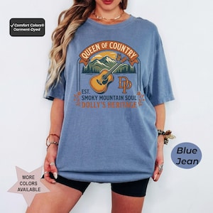 K&ouml;nnte beinhalten: Ein blaues Kurzarm-T-Shirt mit einem Grafikdruck einer akustischen Gitarre, Bergen und dem Text Queen of Country, Est. Smoky Mountain Soul, Dolly's Heritage und DP. Das T-Shirt hat einen Rundhalsausschnitt und eine lockere, weite Passform.