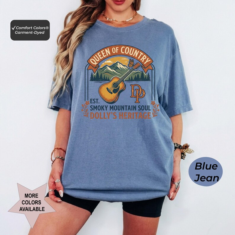 K&ouml;nnte beinhalten: Ein blaues Kurzarm-T-Shirt mit einem Grafikdruck einer akustischen Gitarre, Bergen und dem Text Queen of Country, Est. Smoky Mountain Soul, Dolly's Heritage und DP. Das T-Shirt hat einen Rundhalsausschnitt und eine lockere, weite Passform.