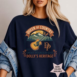 K&ouml;nnte beinhalten: Ein marineblaues T-Shirt mit einem Grafikdruck, der eine akustische Gitarre, Berge und einen Schmetterling zeigt. Der Text lautet QUEEN OF COUNTRY, DP, EST. SMOKY MOUNTAIN SOUL und DOLLY'S HERITAGE. Ein sternf&ouml;rmiges Symbol zeigt MORE COLORS AVAILABLE.