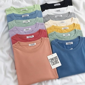 K&ouml;nnte beinhalten: Eine Sammlung von T-Shirts mit Rundhalsausschnitt in verschiedenen Unifarben, darunter Pfirsich, Blau, Gr&uuml;n, Gelb, Lavendel, Grau, Wei&szlig; und Schwarz. Jedes Shirt hat ein wei&szlig;es Innenetikett mit der Aufschrift COMFORT und ein wei&szlig;es Anh&auml;ngeetikett COMFORT COLORS.