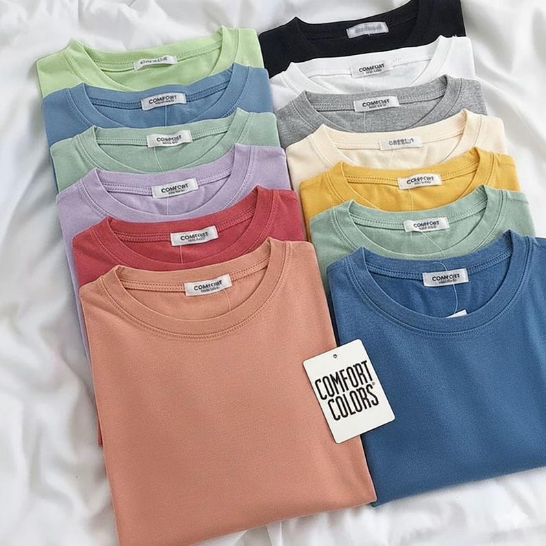 K&ouml;nnte beinhalten: Eine Sammlung von T-Shirts mit Rundhalsausschnitt in verschiedenen Unifarben, darunter Pfirsich, Blau, Gr&uuml;n, Gelb, Lavendel, Grau, Wei&szlig; und Schwarz. Jedes Shirt hat ein wei&szlig;es Innenetikett mit der Aufschrift COMFORT und ein wei&szlig;es Anh&auml;ngeetikett COMFORT COLORS.