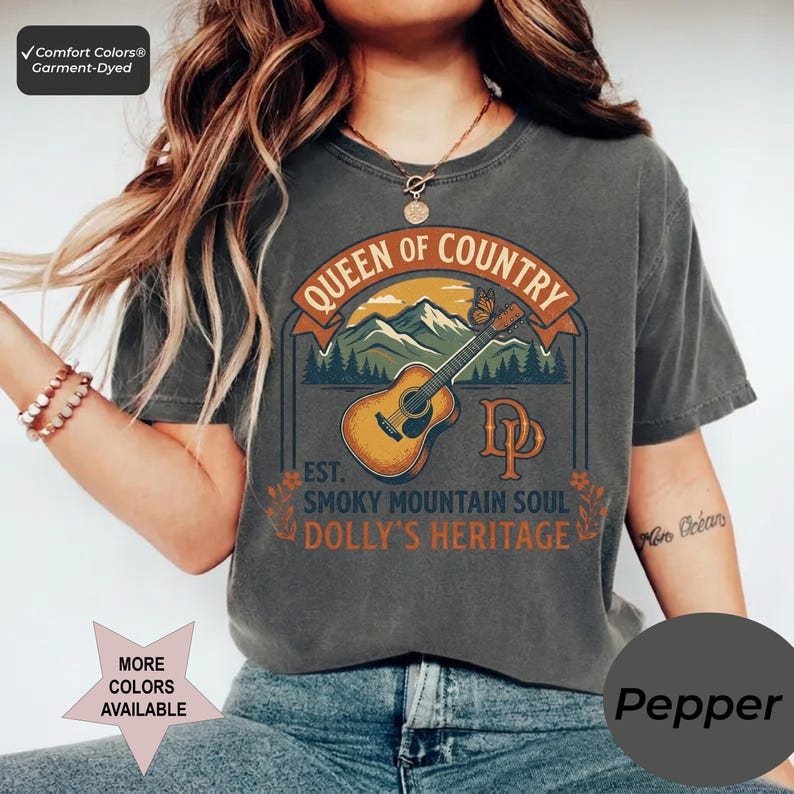 K&ouml;nnte beinhalten: Ein anthrazitfarbenes Kurzarm-T-Shirt mit einem Grafikdruck einer akustischen Gitarre, Bergen und einem Schmetterling. Der Text lautet QUEEN OF COUNTRY, EST. SMOKY MOUNTAIN SOUL, DOLLY'S HERITAGE und DP. Das Shirt hat ein Comfort Colors Etikett.