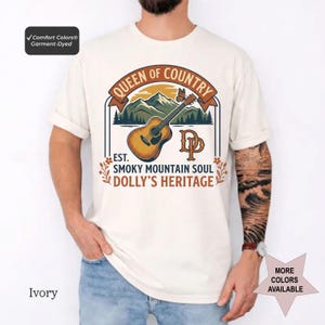 K&ouml;nnte beinhalten: Ein elfenbeinfarbenes Kurzarm-T-Shirt mit einem Grafikdruck, der eine akustische Gitarre, Berge und einen Schmetterling zeigt. Der Text lautet QUEEN OF COUNTRY, EST. SMOKY MOUNTAIN SOUL, DOLLY'S HERITAGE und DP. Ein Comfort Colors Etikett ist sichtbar.