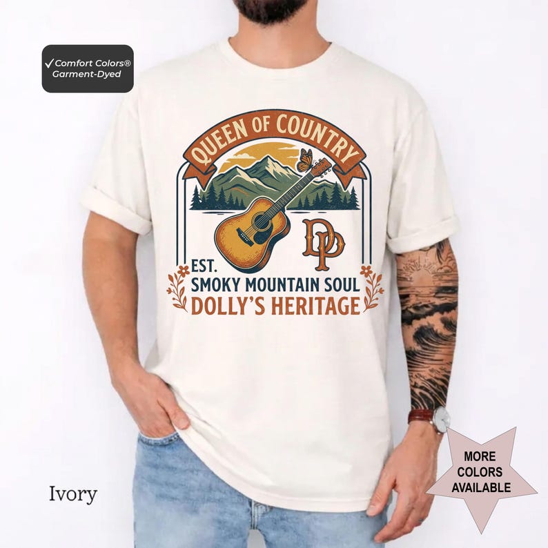 K&ouml;nnte beinhalten: Ein elfenbeinfarbenes Kurzarm-T-Shirt mit einem Grafikdruck, der eine akustische Gitarre, Berge und einen Schmetterling zeigt. Der Text lautet QUEEN OF COUNTRY, EST. SMOKY MOUNTAIN SOUL, DOLLY'S HERITAGE und DP. Ein Comfort Colors Etikett ist sichtbar.