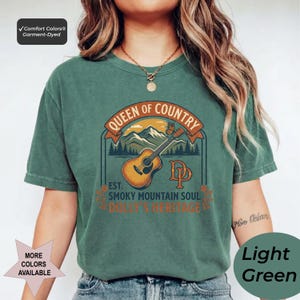 K&ouml;nnte beinhalten: Ein dunkelgr&uuml;nes Kurzarm-T-Shirt mit einem Grafikdruck einer akustischen Gitarre, Bergen und einem Schmetterling. Der Text lautet QUEEN OF COUNTRY, EST. SMOKY MOUNTAIN SOUL, DOLLY'S HERITAGE und DP. Ein Comfort Colors Garment-Dyed Etikett ist sichtbar.