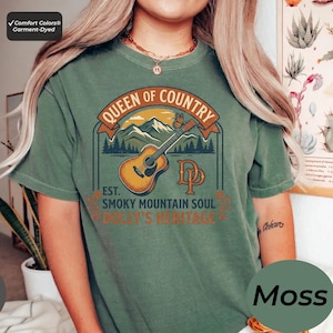 K&ouml;nnte beinhalten: Ein moosgr&uuml;nes Kurzarm-T-Shirt mit einem Grafikdruck mit dem Text Queen of Country, Est. Smoky Mountain Soul und Dolly's Heritage. Das Design zeigt eine braune Akustikgitarre, eine Bergkette, einen Schmetterling und die Buchstaben DP.