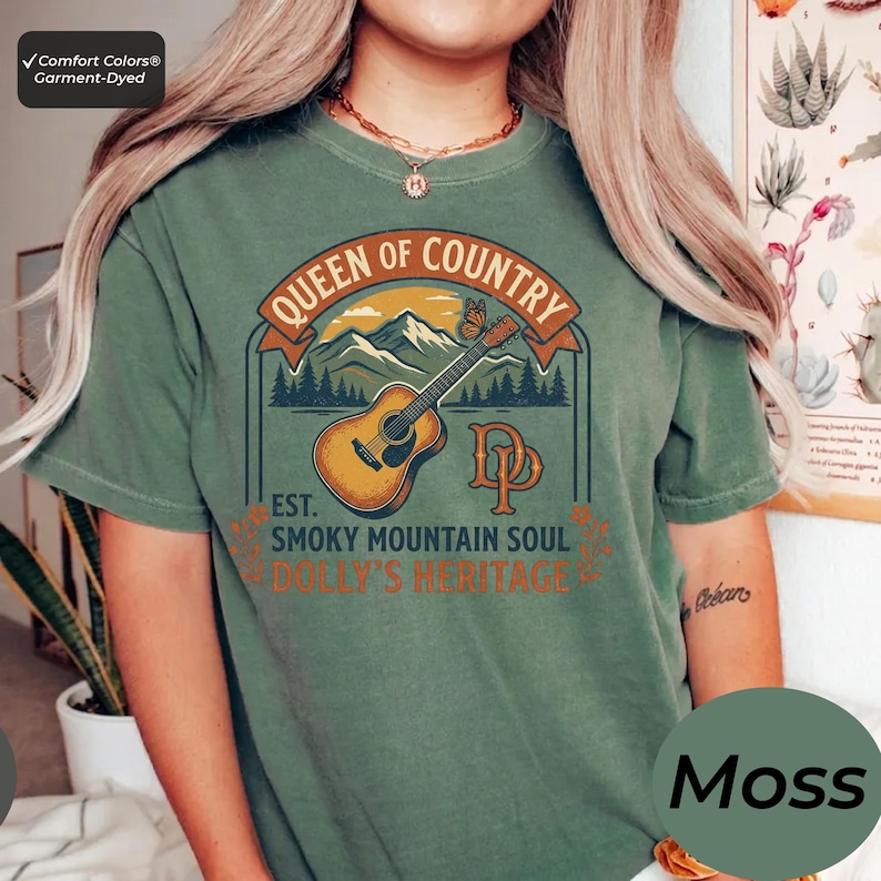 K&ouml;nnte beinhalten: Ein moosgr&uuml;nes Kurzarm-T-Shirt mit einem Grafikdruck mit dem Text Queen of Country, Est. Smoky Mountain Soul und Dolly's Heritage. Das Design zeigt eine braune Akustikgitarre, eine Bergkette, einen Schmetterling und die Buchstaben DP.