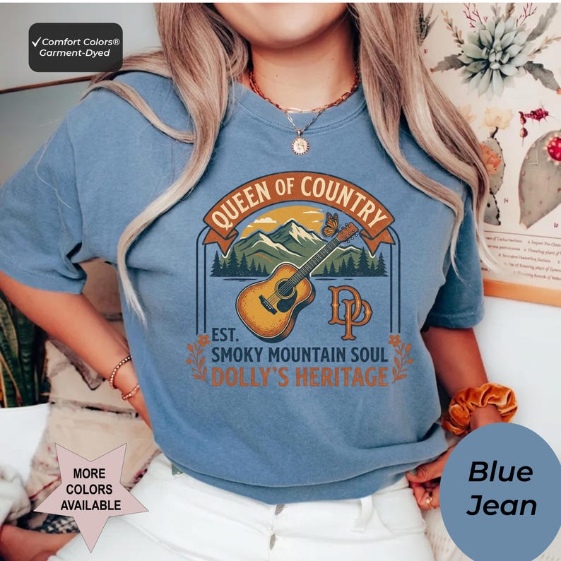 K&ouml;nnte beinhalten: Ein kurz&auml;rmeliges T-Shirt in Jeansblau mit einem Grafikdruck, der eine akustische Gitarre, Berge und einen Schmetterling zeigt. Der Text lautet Queen of Country, Est. Smoky Mountain Soul, Dolly's Heritage sowie die Initialen DP in Orange und Braun.