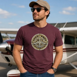 May include: A maroon short-sleeved t-shirt featuring a circular graphic on the chest. The graphic displays a vintage-style compass rose overlaid with a map pattern and a silhouette of a small airplane in the center, with cardinal directions.