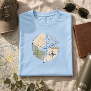 May include: A light blue t-shirt featuring a circular graphic print of a topographic map, a compass rose, and various navigational lines. The design incorporates muted shades of tan, olive green, and dark blue, centred on the chest.