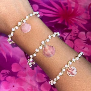 May include: Three bracelets featuring gold-toned wire wrapped with small white beads. Each bracelet includes a single dangling seashell charm in varying shades of pink and white, showcasing natural ridges and textures on the shell surfaces.