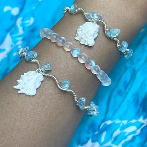 May include: Three bracelets worn together. Two bracelets feature silver-toned wire wrapped with small, light blue, translucent, irregular stones and a white, carved seashell charm. The middle bracelet consists of round, iridescent, frosted beads.