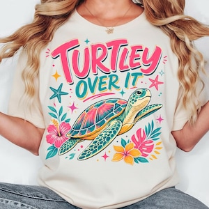 May include: A cream-colored t-shirt featuring a colorful graphic of a sea turtle swimming among tropical flowers and starfish. The text "TURTLEY OVER IT" is printed in large, bold pink and teal letters above the turtle illustration.