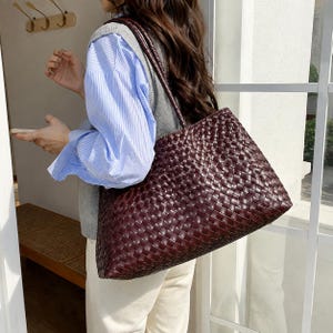 May include: A dark brown tote bag featuring a woven leather-like exterior pattern. The bag has a rectangular shape with a wide base and two long, thin shoulder straps attached to the top. The surface has a glossy finish and a grid-like texture.
