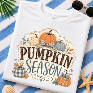 May include: A white t-shirt featuring a graphic of various pumpkins in orange, blue, and cream colors. The design includes a blue plaid bow and the text "PUMPKIN SEASON" in dark brown and blue lettering, surrounded by small leaves and heart accents.
