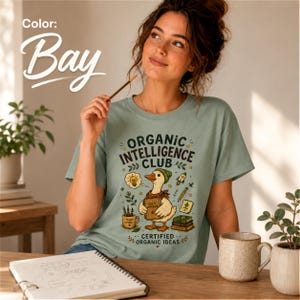 May include: A bay-colored t-shirt featuring a graphic of a duck wearing a beanie and scarf, holding a notebook. The text reads "ORGANIC INTELLIGENCE CLUB" and "CERTIFIED ORGANIC IDEAS," surrounded by illustrations of a lightbulb, rocket, and books.