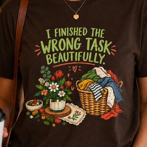 May include: A dark brown short-sleeve t-shirt featuring a graphic print with the text "I FINISHED THE WRONG TASK BEAUTIFULLY." The design includes a colourful bouquet of flowers in a white vase and a woven basket filled with folded laundry items.