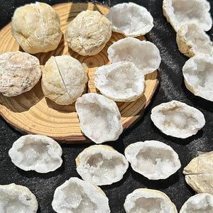 May include: A collection of round, beige-colored geode rocks, some intact and others split open to reveal hollow interiors filled with sparkling, translucent white quartz crystals. Several of these mineral specimens are arranged on a circular wooden slice.