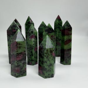 May include: A collection of nine polished zoisite crystal towers, each featuring a hexagonal prism shape with a pointed top. The stones display a mottled pattern of deep forest green, vibrant magenta-red, and dark black mineral inclusions.