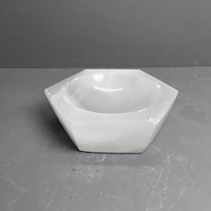 May include: A white, translucent, hexagonal bowl carved from selenite. The object features a smooth, polished surface with a shallow, rounded interior basin and flat, angled exterior sides that form a distinct six-sided geometric shape.