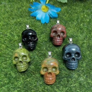 May include: Five skull-shaped pendants carved from various stones, including black, red, green, and grey varieties. Each pendant features a silver-toned metal bail at the top for attachment, showcasing detailed facial features like eye sockets and teeth.