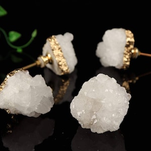May include: A pair of stud earrings featuring raw, white druzy quartz crystals. Each crystal has a textured, gold-toned metallic edge and a standard post backing. The stones have a jagged, natural appearance with a sparkling, crystalline surface.