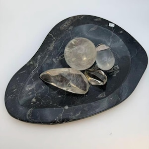 May include: A teardrop-shaped black stone tray with white veining and fossil inclusions. Resting on the tray are four clear, polished stones: one large sphere, one smooth oval, one elongated irregular piece, and one small faceted crystal.