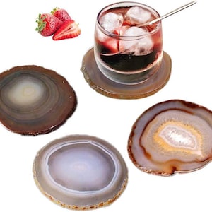 May include: A set of four circular agate stone coasters with natural banded patterns in shades of brown, gray, and white. One coaster supports a clear glass filled with a dark red beverage, ice cubes, and a metal stirring spoon, near fresh strawberries.