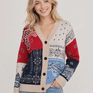May include: A long-sleeve V-neck cardigan featuring a patchwork design of red, navy blue, light blue, and cream panels. The knit garment displays intricate bandana-style patterns in black and white, finished with a beige ribbed trim and four black buttons.