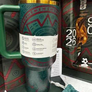 May include: A dark green stainless steel tumbler with a handle and a gold-colored rim, featuring a red and green geometric pattern. A white paper band wraps around the center, displaying product icons and text. The tumbler is positioned in front of a FIFA World Cup 2026 branded box.