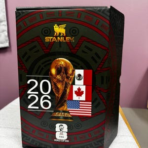 May include: A black rectangular product box featuring the Stanley logo in gold, the text 2026, and a gold FIFA World Cup trophy graphic. The design includes the flags of Mexico, Canada, and the United States, along with the official FIFA World Cup 2026 logo.