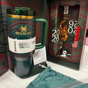 May include: A green Stanley Flowstate Quencher H2.0 tumbler with a 40 oz capacity, featuring a gold rim and a patterned design. It is presented in a box with FIFA World Cup 2026 branding, including a graphic of the trophy and a green fabric pouch.