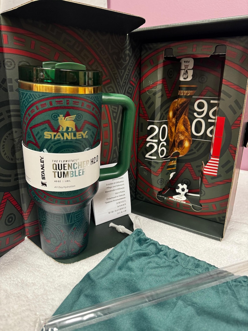 May include: A green Stanley Flowstate Quencher H2.0 tumbler with a 40 oz capacity, featuring a gold rim and a patterned design. It is presented in a box with FIFA World Cup 2026 branding, including a graphic of the trophy and a green fabric pouch.