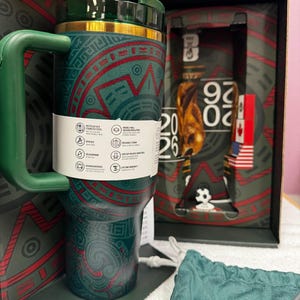 May include: A dark green stainless steel tumbler with a handle and a gold-tone rim, featuring red and green geometric patterns. A white product information band wraps around the center. The tumbler is displayed in a box marked with 2026.
