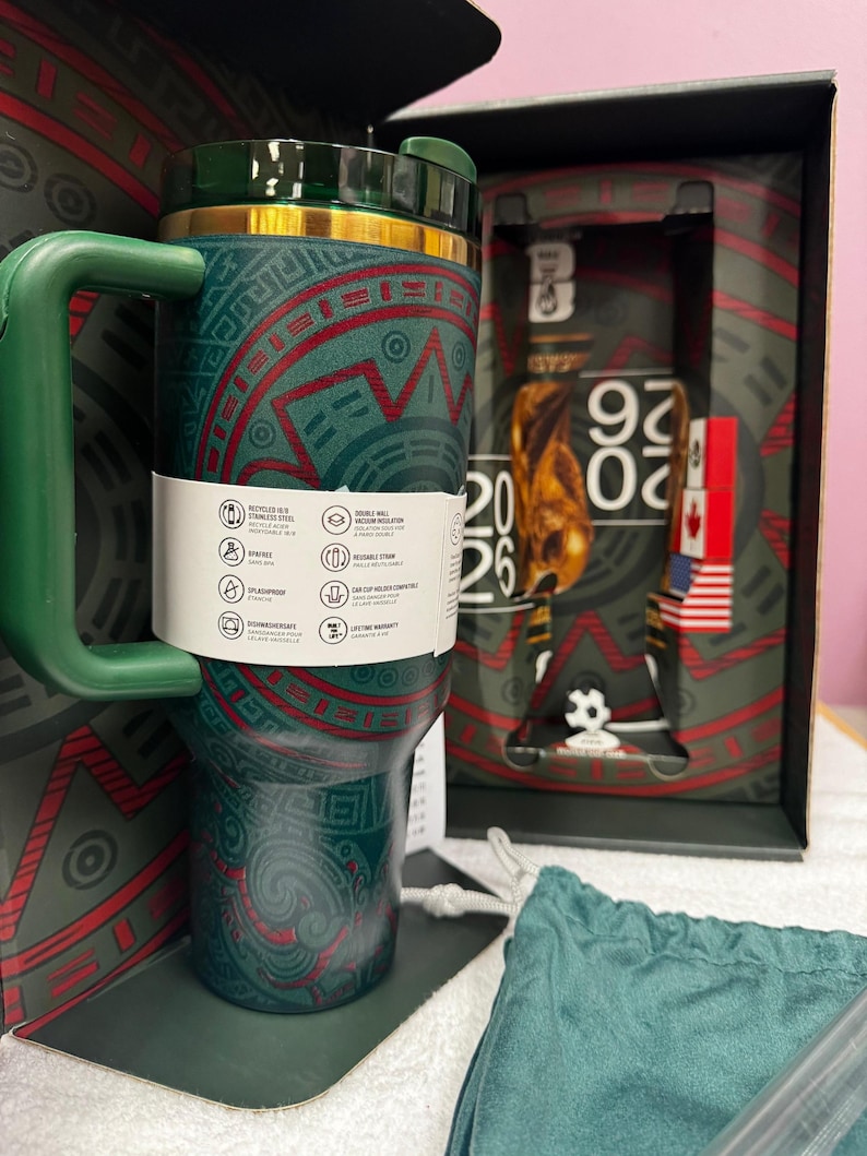 May include: A dark green stainless steel tumbler with a handle and a gold-tone rim, featuring red and green geometric patterns. A white product information band wraps around the center. The tumbler is displayed in a box marked with 2026.