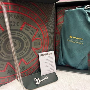 May include: A Stanley and LoveShackFancy branded dark green fabric drawstring bag, a clear plastic straw, and a small soccer ball-shaped straw topper inside a decorative box with a red and dark gray circular pattern and an instruction booklet.