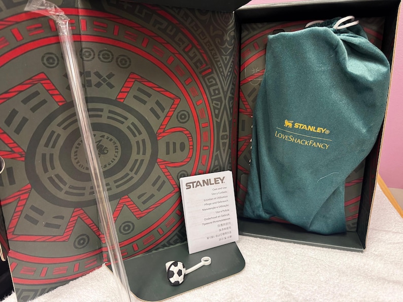 May include: A Stanley and LoveShackFancy branded dark green fabric drawstring bag, a clear plastic straw, and a small soccer ball-shaped straw topper inside a decorative box with a red and dark gray circular pattern and an instruction booklet.