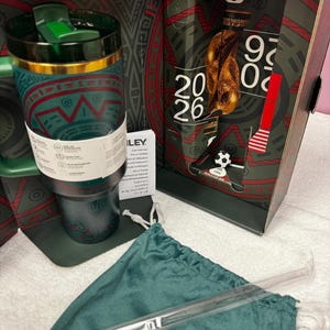 May include: A dark green stainless steel tumbler with a gold rim and a green lid, featuring red and teal geometric patterns. It is presented in a box marked FIFA World Cup 2026, accompanied by a matching green drawstring pouch and a clear straw.