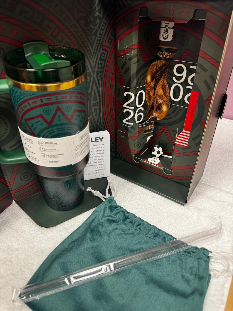 May include: A dark green stainless steel tumbler with a gold rim and a green lid, featuring red and teal geometric patterns. It is presented in a box marked FIFA World Cup 2026, accompanied by a matching green drawstring pouch and a clear straw.
