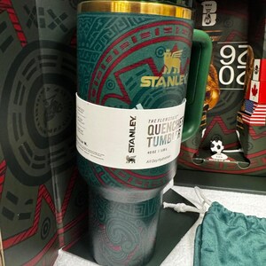 May include: A dark green Stanley Flowstate Quencher tumbler with a 40 oz capacity. The exterior features a gold-colored rim and a patterned design with red accents. A white paper band wraps around the center, displaying the text Stanley, The Flowstate Quencher Tumbler, 40 oz | 1.18 L, and All Day Hydration.