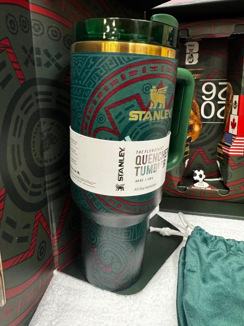 May include: A dark green Stanley Flowstate Quencher tumbler with a 40 oz capacity. The exterior features a gold-colored rim and a patterned design with red accents. A white paper band wraps around the center, displaying the text Stanley, The Flowstate Quencher Tumbler, 40 oz | 1.18 L, and All Day Hydration.