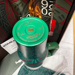 May include: A dark green Stanley tumbler with a 40oz/1.18L capacity, featuring a matching green lid with a gold logo, a side handle, and a patterned body. The tumbler is accompanied by a clear straw, a green fabric pouch, and product packaging.