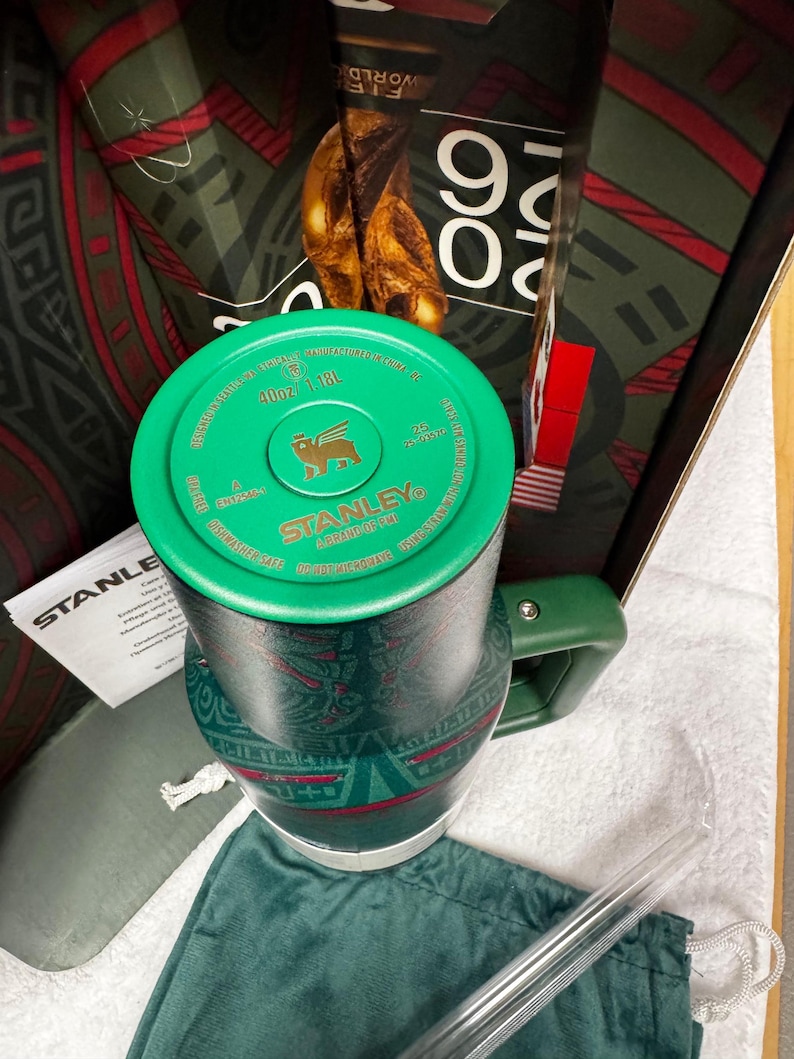May include: A dark green Stanley tumbler with a 40oz/1.18L capacity, featuring a matching green lid with a gold logo, a side handle, and a patterned body. The tumbler is accompanied by a clear straw, a green fabric pouch, and product packaging.