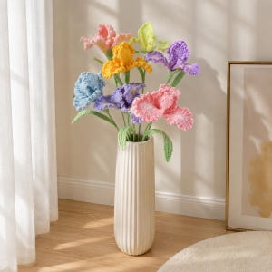 Puede incluir: Un ramo de flores de iris tejidas a ganchillo en tonos rosa, amarillo, azul claro, morado y verde lima, cada una con tallo y hojas verdes, dispuesto en un jarrón cilíndrico alto de cerámica blanca con textura acanalada vertical.
